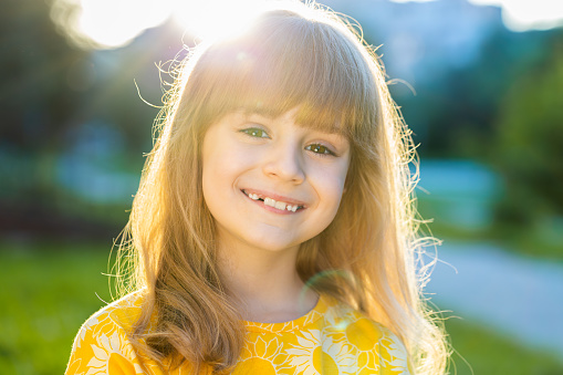 Portrait of happy preteen child kid smiling friendly, glad expression looking at camera dreaming, resting, relaxation feel satisfied good news. Pretty blonde girl on city sunset park street, outdoors