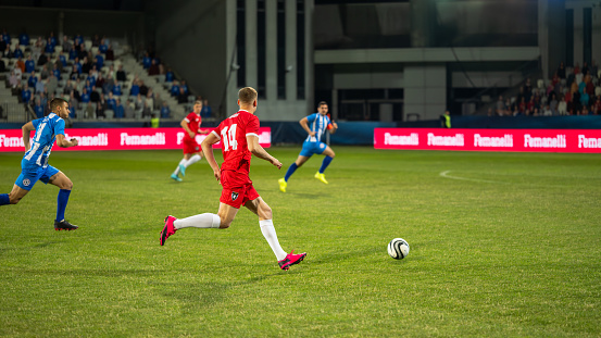 Male soccer players running and leading ball during match on sports field, side view wide shot