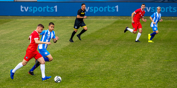 Male soccer players running and leading ball during match on sports field, full length shot