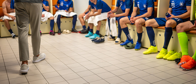 Soccer coach talking with male players team in locker room, legs medium shot