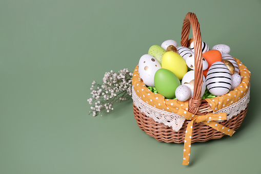 Wicker basket with festively decorated Easter eggs and gypsophila flowers on pale green background. Space for text