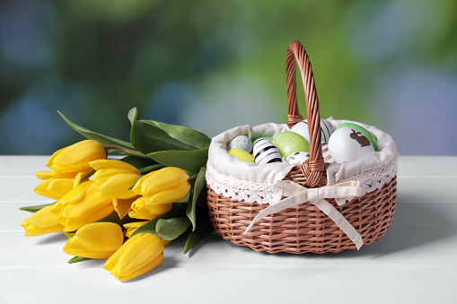 Wicker basket with festively decorated Easter eggs and beautiful tulips on white wooden table