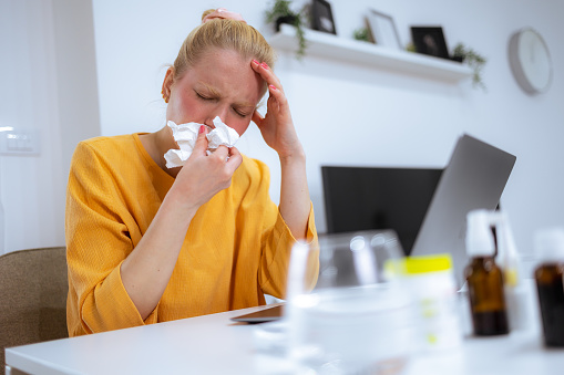 Allergic  woman blowing nose in tissue sit at home office study work on laptop, ill sick  girl got flu caught cold sneeze in tissue having allergy symptoms coughing holding napkin