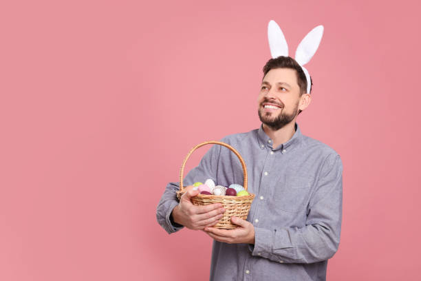 istock Happy man in bunny ears headband holding wicker basket with painted Easter eggs on pink background. Space for text 1777214089