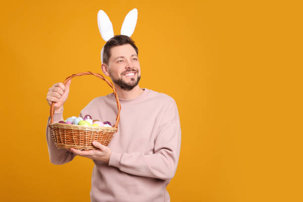 istock Happy man in bunny ears headband holding wicker basket with painted Easter eggs on orange background. Space for text 1777214143