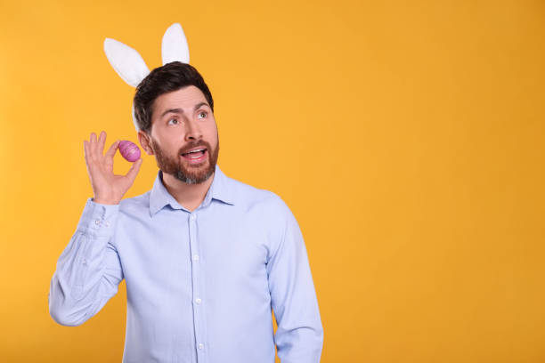 istock Emotional man in cute bunny ears headband holding Easter egg on orange background. Space for text 1777214151