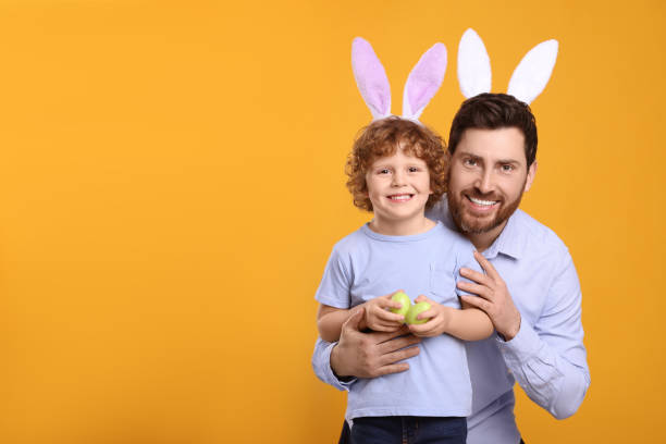 オレンジ色の背景にかわいいウサギの耳のカチューシャを身に着けている幸せな父と息子。イースターエッグを持つ少年、テキスト用のスペース - bunny painting ストックフォトと画像