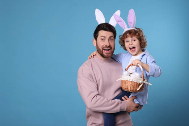 istock Happy father and son in cute bunny ears headbands on light blue background. Boy holding Easter basket with painted eggs, space for text 1777215820
