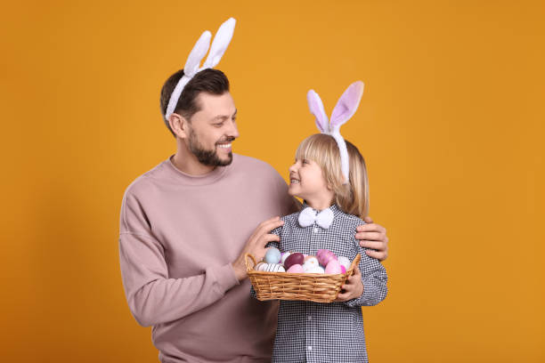 istock Father and son in bunny ears headbands with wicker basket of painted Easter eggs on orange background 1777215821