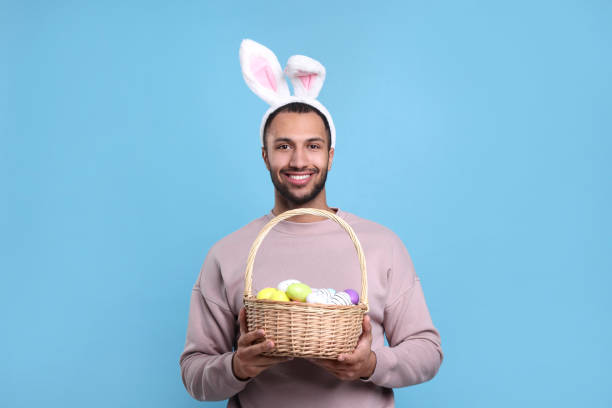 水色の背景にイースターエッグと籐のバスケットを保持しているウサギの耳のヘッドバンドの幸せなアフリカ系アメリカ人の男性 - bunny painting ストックフォトと画像