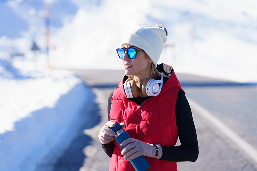 Young inspired female in warm clothes and sunglasses holding thermos bottle while having workout in snowy mountains