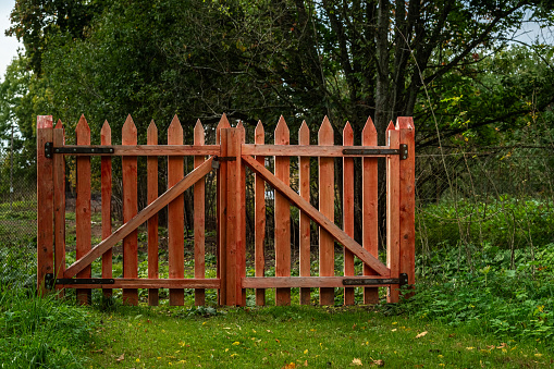 Closed gates made of wooden picket fence, painted red, against a background of green trees.