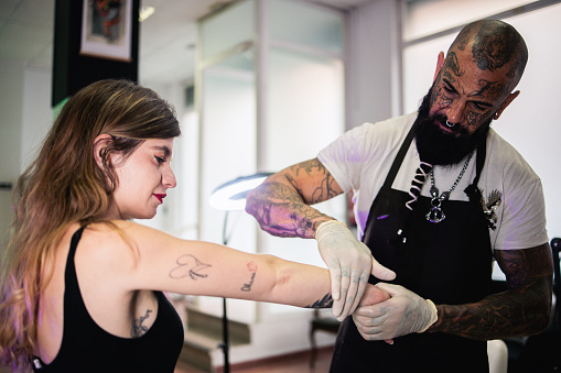 Tattoo artist working at his studio and preparing to do a tattoo on a young woman's arm.