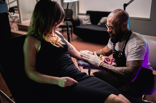 Tattoo artist working at his studio and tattooing a young woman's arm.