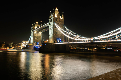 London, England, United Kingdom  - December 17, 2022: Profile shot of the beautifully illuminated Tower Bridge over the Thames River at night with city lights in distance.