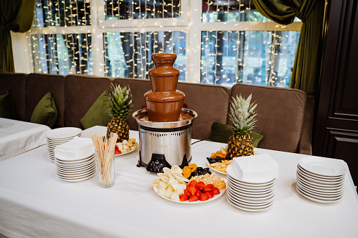 a chocolate fountain stands on a white table with sliced fruit next to it. dessert table. Sweet table on the occasion.