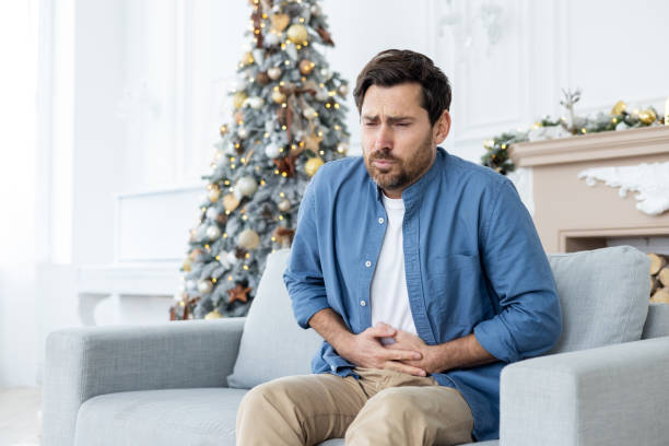 un joven sufre de fuertes dolores abdominales durante las vacaciones de navidad. se sienta en el sofá de su casa cerca del árbol de navidad y se sostiene el cuerpo con las manos, haciendo muecas por los espasmos - christmas eating nausea illness fotografías e imágenes de stock