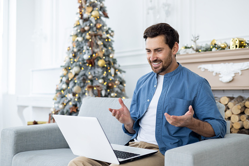 A young man is talking on a video call on a laptop while sitting on the sofa at home ,near the Christmas tree on the eve of the Christmas holidays.