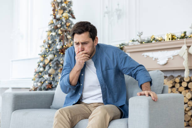 un joven se sienta en el sofá de su casa durante las vacaciones de año nuevo y siente muchas náuseas, se tapa la boca con la mano, quiere vomitar. intoxicación etílica, envenenamiento - christmas eating nausea illness fotografías e imágenes de stock