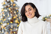 Close-up photo of a portrait of a young beautiful brunette woman in a white sweater at home against the background of a Christmas tree. He looks at the camera with a smile