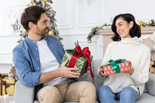 Young happy couple in love, family, man and woman sitting on sofa at home and greeting each other with Christmas presents and surprises for New Year holidays.