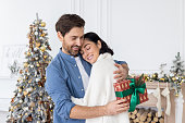 A young couple, a family, a man and a woman celebrate Christmas and winter holidays together at home, hug by the Christmas tree, exchange gifts, congratulate each other