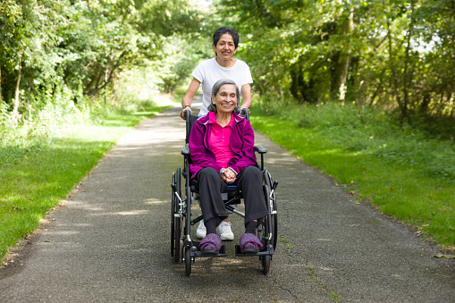 Asian Indian woman pushing her elderly mother in a wheelchair outdoors in summer, UK. May also depict a carer, care in the community