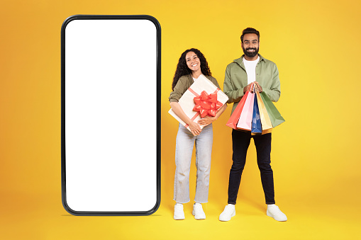 Shopping App. Cheerful Millennial Arab Customers Couple Near Big Cellphone With Empty Screen On Yellow Studio Background, Holding Gift Box And Colorful Shopper Bags. Mockup, Sales Offer Ad. Collage