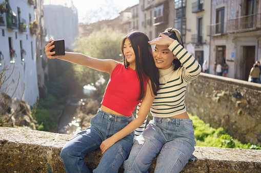 Happy young female friends in casual clothes smiling and taking selfie on smartphone while sitting on stone bench in Granada