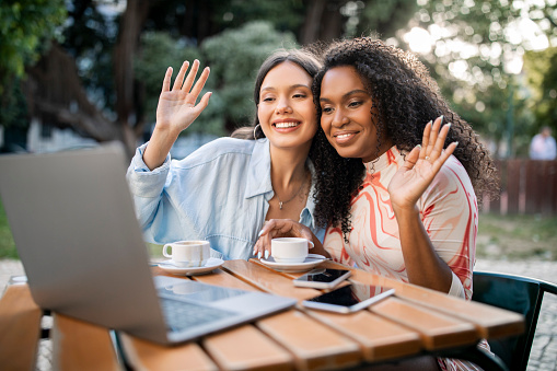 Two Happy Female Friends Making Video Call On Laptop Outdoors While Sitting On Summer Terrace At Cafe, Cheerful Multiethnic Besties Waving Hands At Computer Web Camera, Enjoying Online Communication