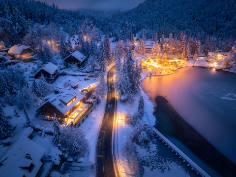Aerial view of fairy town in snow, road, forest, Jasna lake and houses with lights at night in winter. Top view of mountain village, illumination, snowy pine trees at dusk in Kranjska Gora, Slovenia