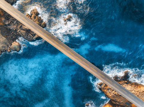 Aerial view of road, sea with waves and stones at sunset in Lofoten Islands, Norway. Landscape with beautiful bridge, transparent blue water, rocks. Top view from drone of highway in summer. Travel