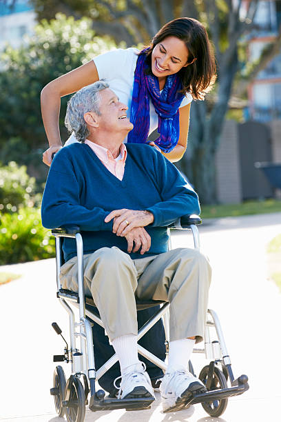 Adult Daughter pushing her father in a wheelchair outdoors Adult Daughter Pushing Senior Father In Wheelchair Looking At Each Other Talking a helping hand photos stock pictures, royalty-free photos & images