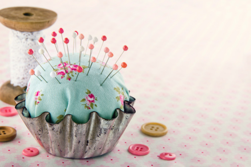 Green handmade floral pincushion in an old metal cupcake with buttons and spools of thread and lace, sewing concept background