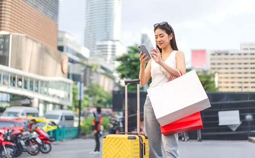 People city lifestyles travel with buying shopping consumerism. Young adult asian woman using smartphone with luggage. Happy smile face standing at outdoors.