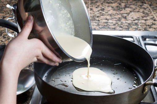 Horizontal photo of focus on pancake batter coming out of stainless steel bowl and into hot frying pan