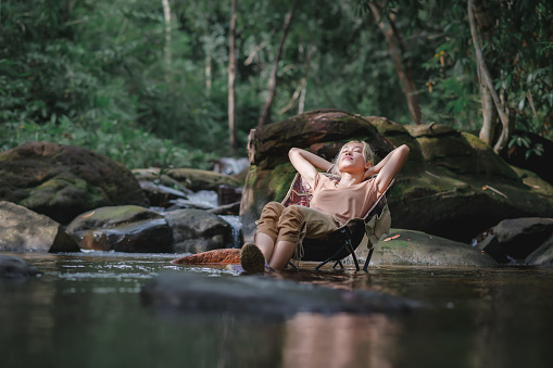 Young woman sitting on a camping chair in the stream and reclining to relax, Admiring the surrounding natural atmosphere with weak sunlight. Healthy Lifestyle by Nature Therapy.