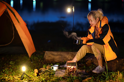 Female traveler or Backpacker sits by the camping stove near her tent, Boiling the water and easily cooking ready-to-eat food in a moment of relaxation beneath the blue sky and atmosphere of nature.