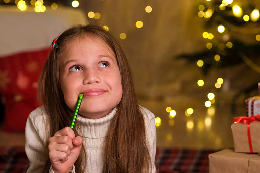 Cute child girl is writing letter to Santa Claus in decorated room for Christmas. Close up kid making wishes near gifts on background of Christmas tree. Holidays and celebrations concept