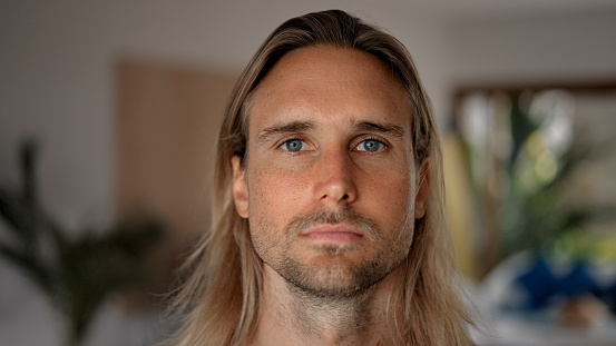 Unhappy male person face home portrait. Blue eyes looking at camera close up. Sad bristle surfer man long hair. Upset shy guy. Kind casual people. Young adult hippie inside house. Hipster hairstyle.