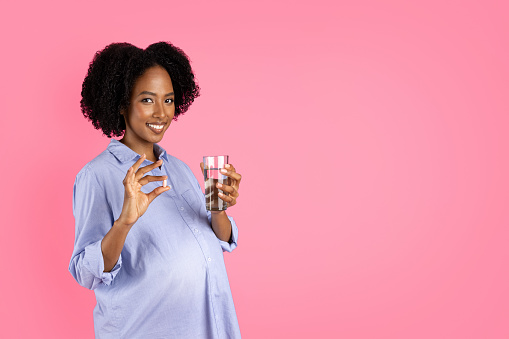 Happy millennial pregnant black lady with big belly show pill capsule and glass of water, isolated on pink background, studio. Recommendation of vitamins, minerals, health care and medicine