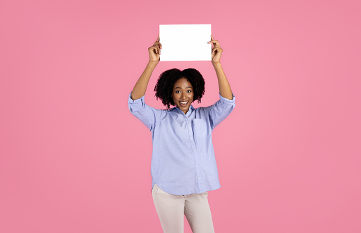 Smiling young pregnant african american curly lady in casual with big belly hold banner with blank space, isolated on pink studio background. Expect baby, recommendation, healthcare, ad and offer