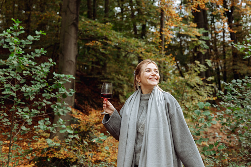 Hello November. smiling fashionable 40-year-old woman in a gray coat with a glass of wine enjoys autumn outdoors in the forest. High quality photo