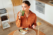 Young caucasian lady having dinner, drinking a glass of water and tasting a caesar salad, sitting in kitchen, above view