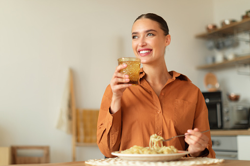 Happy young woman holding glass of water while eating homemade spaghetti, sitting at table in kitchen interior and looking aside, free space