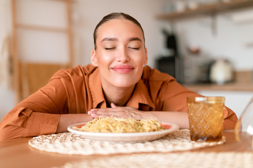 Italian cuisine. Happy satisfied european woman smelling fresh spaghetti on plate, enjoying tasty homemade pasta, sitting in kitchen interior