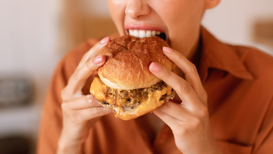 Hungry caucasian woman enjoy eating tasty hamburger, biting home-prepared or delivered junk food meal, closeup, cropped, panorama. Junk food concept