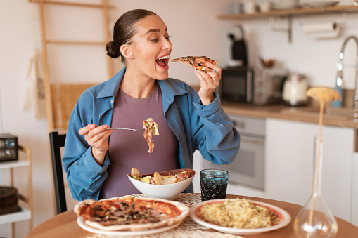 Expectant delights. Hungry pregnant woman eating pizza and salad, having desire for junk food, sitting at table in kitchen interior. Cheat meal, gluttony and fastfood concept
