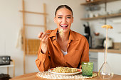 Happy young european lady eating homemade Italian pasta, enjoying tasty lunch, sitting in kitchen interior, copy space