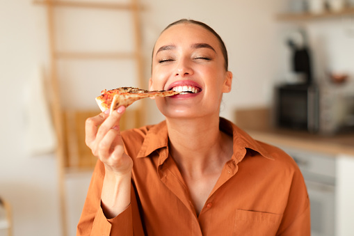 Contented young caucasian woman enjoying her kitchen creation, biting slice of homemade pizza, having dinner, sitting at table in kitchen, free space, closeup